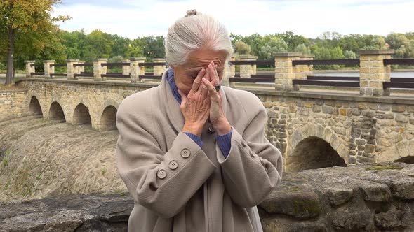 An Elderly Woman Leans Against the Stone Barrier of a Bridge, Looks Upset