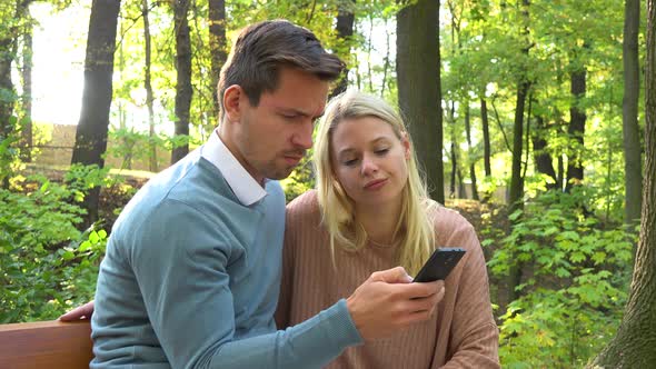 A Young Attractive Couple Looks at a Smartphone on a Bench in a Park on a Sunny Day - Closeup