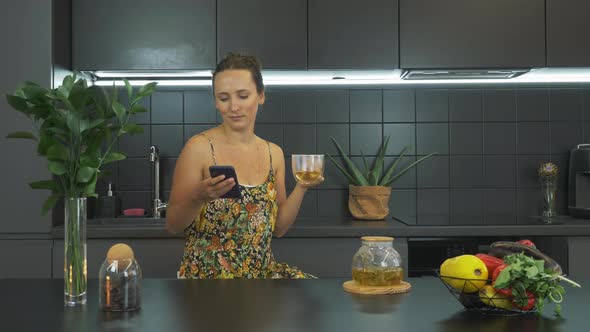 Happy smiling woman is using smartphone and drinking herbal tea, spending free time at home