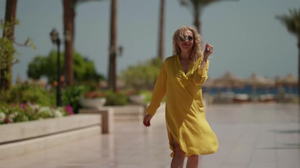 Curly Blonde in Sunglasses and a Yellow Fluttering Dress Poses on a Sunny Day