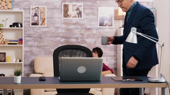 Old Man Holding a Cup of Coffee While Sitting at Desk