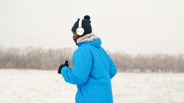 Black man at jacket, hat and headphones is jogging in winter outdoors, side view