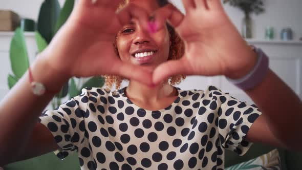 Young African American Woman Shows Gesture of Love Sits on Sofa at Home