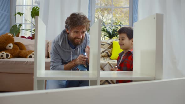 Delighted Little Boy in Casual Clothes Laughing and Helping Father Assembling Furniture at Home