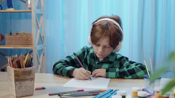 Little Schoolboy Sitting at Table in Room Drawing with Pencil in Album and Listens to Music in Big