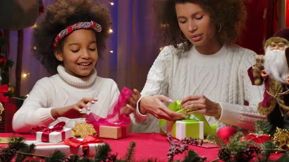 African American Mom and Little Daughter in White Sweaters Decorate Gift Boxes and Tie Holiday Bows