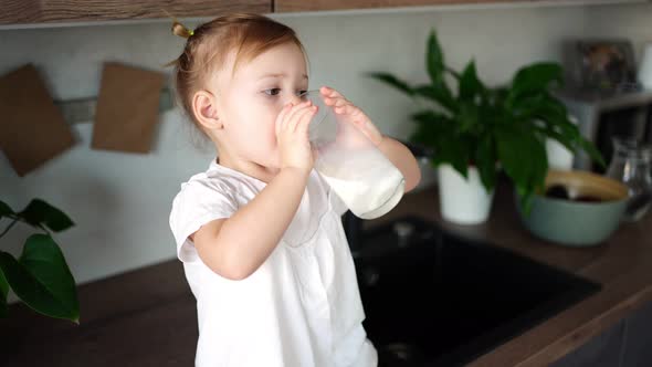 Happy Baby Girl Sitting at the Table in the Kitchen and Drinking Milk