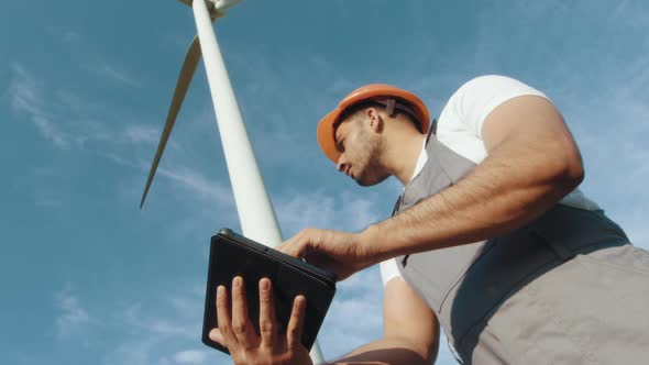 Indian Engineer in Helmet and Grey Overalls Standing Among Windmill Farm with