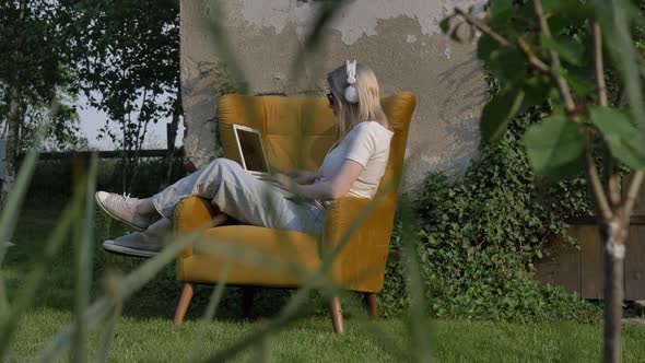 Woman sitting in an armchair using laptop in a garden with wall and doors on background