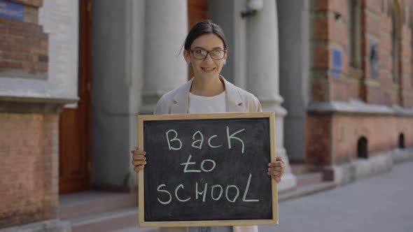 Female Teacher Standing Outdoor Looking Down and at Camera Holding Board Text Back School