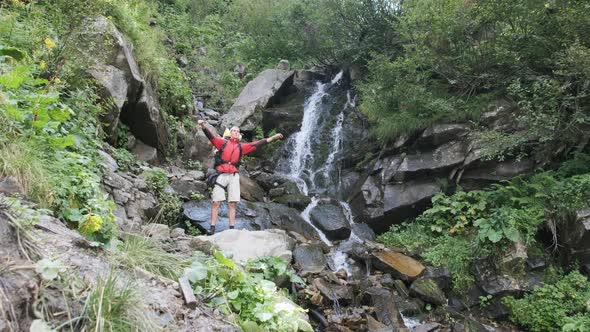 Tourist with Backpack Came To Mountain Waterfall and Spreading Arms To Sides