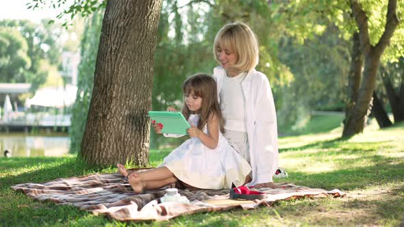 Curios Little Daughter and Beautiful Young Mother Surfing Internet on Tablet Sitting on Blanket in