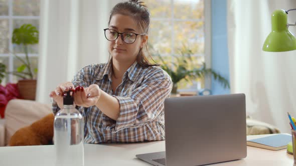 Woman Cleaning Hands with Sanitizer Gel Sitting at Table and Working on Laptop in Home Office