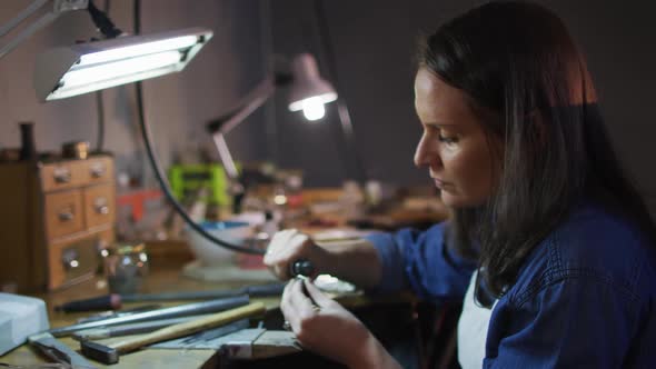 Focused caucasian female jeweller sitting at desk, making jewelry in workshop