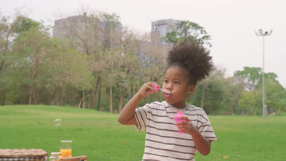 Happy little african american curly hair girl blowing soap bubbles playing alone in the park