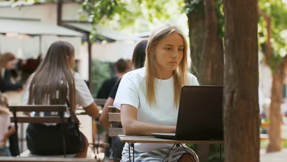 Young Lady is Sitting at Table in a Street Cafe and Typing on Her Laptop Computer