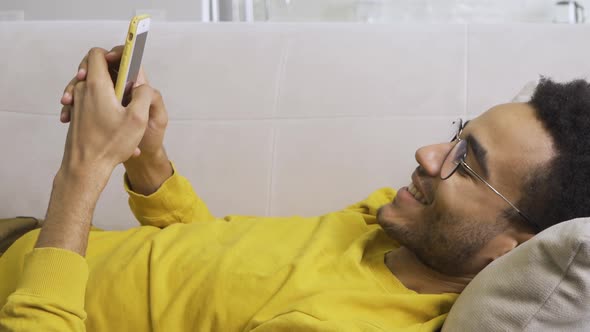 African American Man Lies Resting on the Couch at Home and Holds a Smartphone