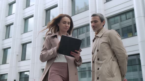 A beautiful girl with a tablet approaches the head of a man standing near office buildings