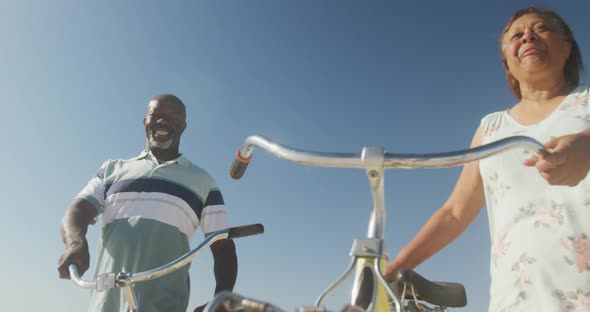 Smiling senior african american couple walking with bicycles on sunny beach