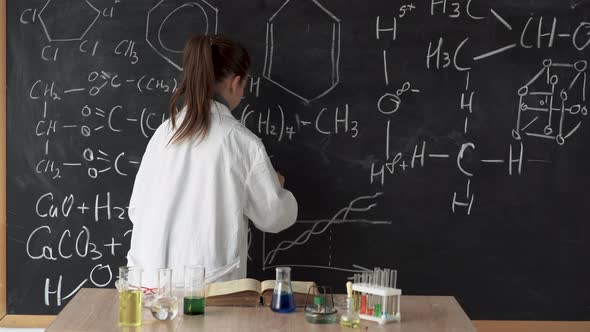 A Little Scientist Girl in a White Coat Writes a Formula on a Blackboard with Chalk
