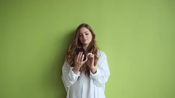 Girl With Long Hair Stands Against Light Green Wall With Mobile Phone In Her Hands