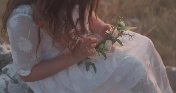 Woman in a white dress sits on a mountain and makes a wreath of flowers at sunset