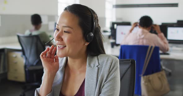 Happy asian businesswoman sitting at desk using computer and talking on headset