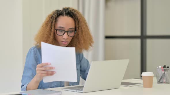 African Woman Reading Documents While Working on Laptop