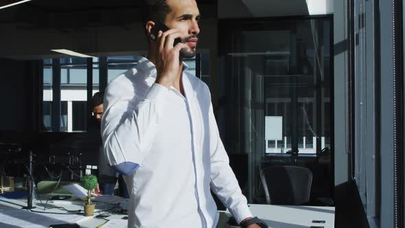 Mixed race businessman standing in sunny office talking on smartphone and looking out of window