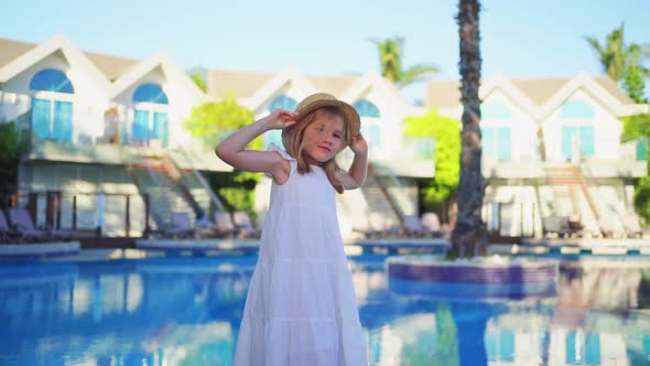 Cute Little Girl in a White Dress and Straw Hat By the Pool Near the Villa