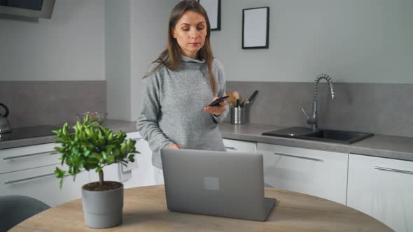 Woman with Smartphone in Hand Standing in Home Cozy Kitchen Makes Herself Coffee and Use Laptop Do