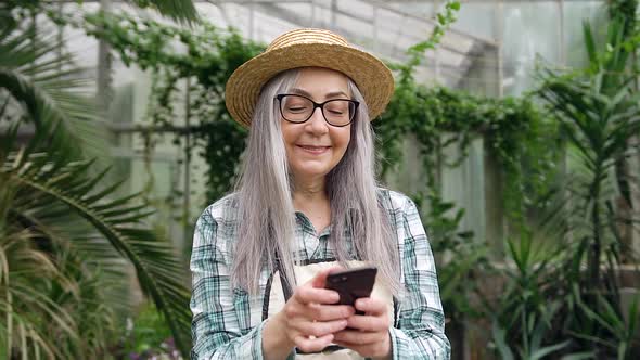 Senior Woman in Hat which Using Smartphone in Beautiful Orangery