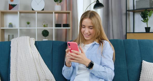 Blondie in Casual Clothes Sitting on Soft Sofa in Living-Room and Writing Message on Phone