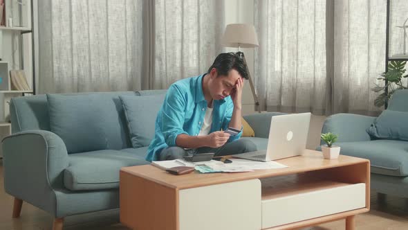 Asian Man With A Laptop Holding And Looking At Credit Card Then Having Headache