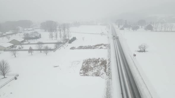 Drone view in snow storm over homes, farm fields and highway in a rural valley, alt