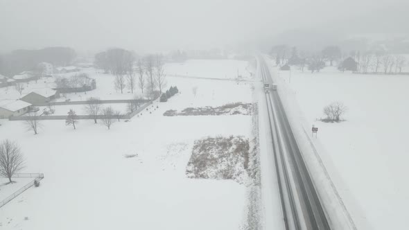 Drone view in snow storm over homes, farm fields and highway in a rural valley,