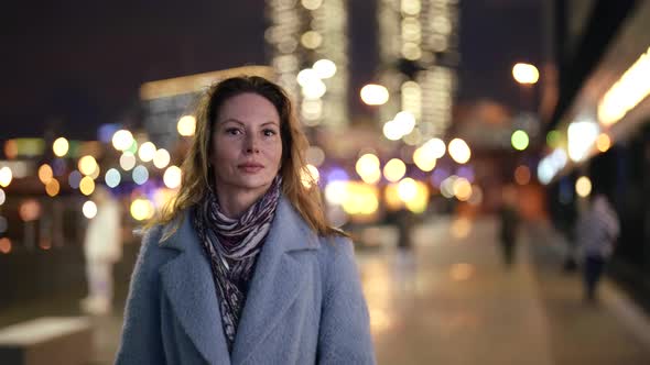 a Blonde with Dark Eyes in a Light Coat and Scarf Walks Along an Evening Street Against the