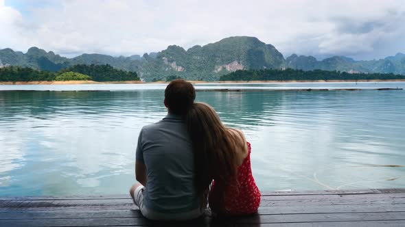 Happy Romantic Couple Sitting Together on Wooden Bridge on Green Mountains Lake