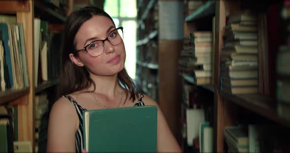 Girl in Glasses with Book in Hands Smiling Lightly at Camera in Library