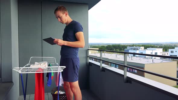 A Young Man Takes Down Clothes From the Drying Rack on the Balcony and Works on the Tablet