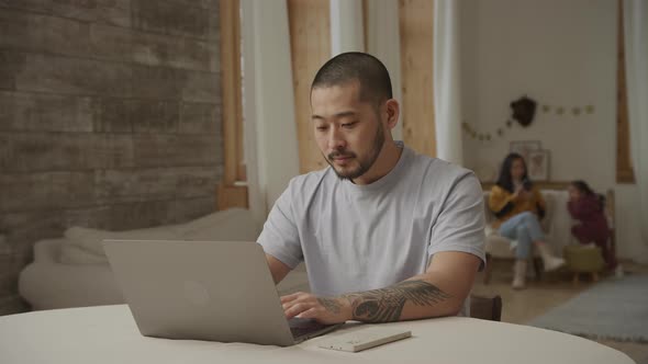 A Happy Father Working on a Laptop While His Family is Playing on the Background Indoors