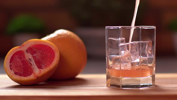 Fresh Grapefruit Juice Pouring Into a Transparent Glass with Ice. Sliced Grapefruits on a Table