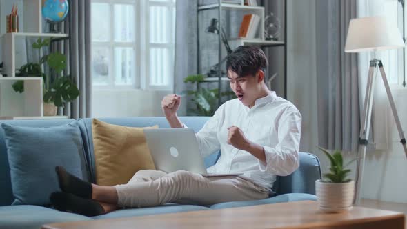 Happy Asian Man Celebrating While Lying On Sofa And Using Laptop Computer In The Living Room