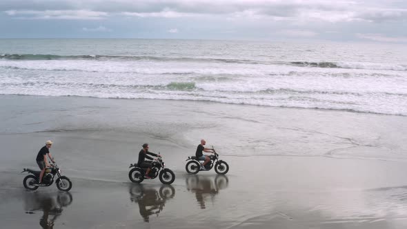 Three Handsome Hipster Men Riding Modern Custom Motorcycle Cafe Racer on the Black Sand Beach