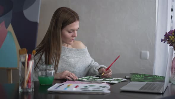 Portrait of Slim Beautiful Artist Painting Picture Looking at Laptop Screen Sitting at Table Indoors