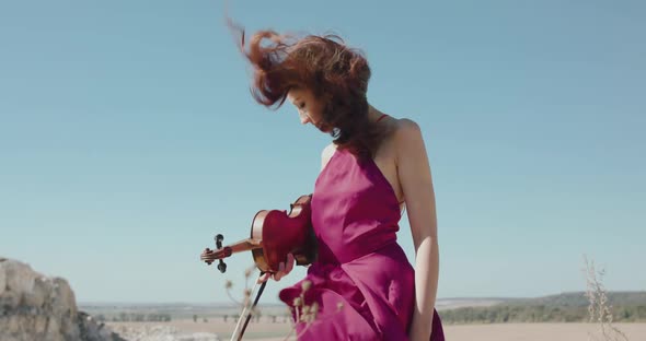 Girl with Blowing Long Hair and Windy Dress Stands with Violin in Hand on Cliff