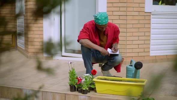 Wide Shot of Concentrated Male African American Gardener Sitting in Squat Writing with Pen on Paper