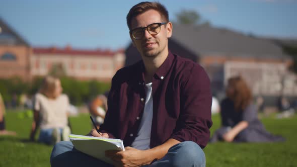 Portrait of Handsome Hipster Guy Student Sitting on Campus Lawn with Book Looking at Camera