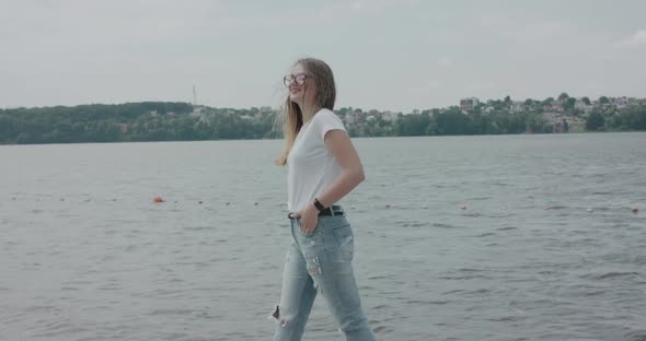 Joyful Girl in Glasses Walking at Riverside and Posing at Camera in Windy Day