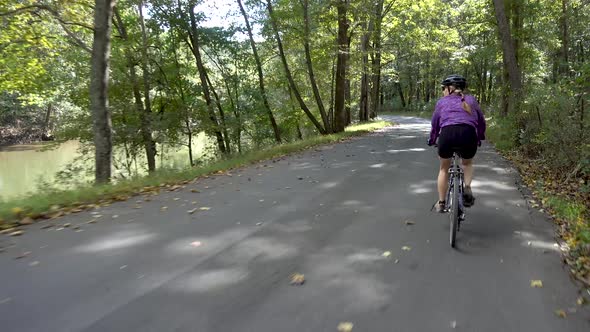 Woman biking on a shady road following along a river.